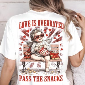 May include: White t-shirt with a graphic featuring a cherub wearing heart-shaped sunglasses, holding popcorn, and sitting on a bench. The text "LOVE IS OVERRATED" and "PASS THE SNACKS" are printed in red.