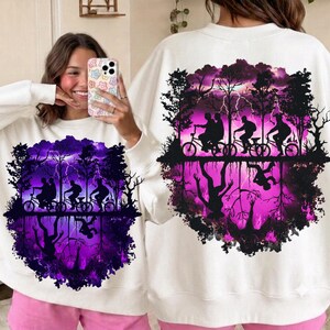 Puede incluir: Una sudadera blanca de manga larga con un estampado gráfico de tres siluetas montando en bicicleta contra un cielo púrpura y rosa lleno de rayos, con un reflejo invertido de la escena debajo, incluyendo árboles y la silueta de una criatura oscura.