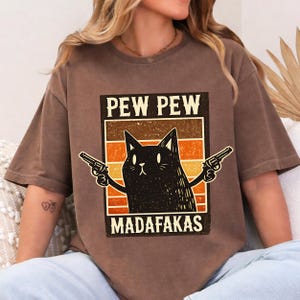 May include: Brown t-shirt featuring a black cat graphic holding two guns, with the text "PEW PEW MADAFAKAS" in a retro design. The cat has a stern expression. The shirt has a relaxed fit.