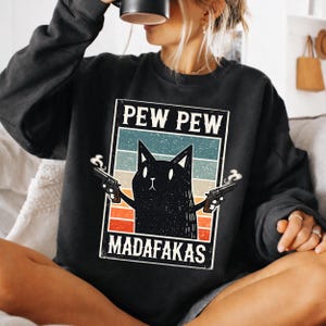 Puede incluir: Sudadera negra con un gráfico de un gato negro sosteniendo dos pistolas, con el texto "PEW PEW MADAFAKAS" en un marco rectangular. El diseño tiene un esquema de color retro con rayas azules, naranjas y blancas.