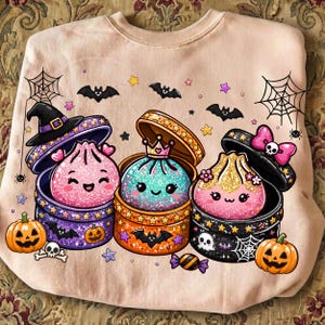 May include: A light peach-colored sweatshirt featuring a graphic print of three glittery, smiling dumplings in round containers. The design includes Halloween elements like pumpkins, bats, spiderwebs, a witch hat, a crown, and a skull bow.