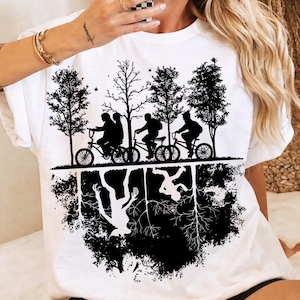 Peut inclure: T-shirt blanc avec un motif noir et blanc représentant quatre personnes à vélo sous des arbres. En dessous, une image miroir montre un monstre et des racines. Le design évoque l'aventure et le mystère.