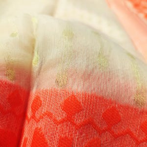 May include: Close-up of a sheer fabric with a cream-colored base. The fabric has gold flecks and a vibrant orange border with a repeating floral pattern. The material is lightweight and appears to be folded.