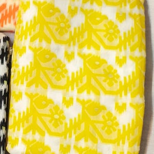 May include: Close-up of a folded fabric with a repeating pattern. The fabric features a bright yellow floral design against a white background. Other fabrics with different patterns are partially visible.