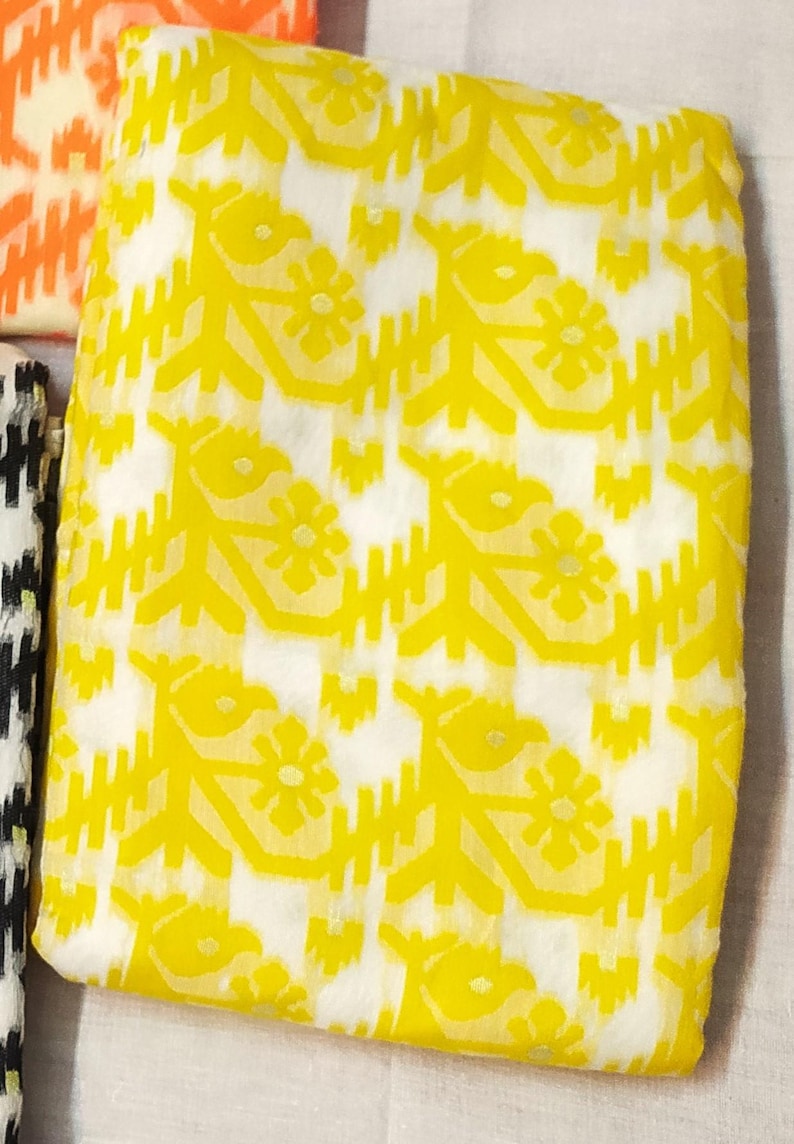 May include: Close-up of a folded fabric with a repeating pattern. The fabric features a bright yellow floral design against a white background. Other fabrics with different patterns are partially visible.