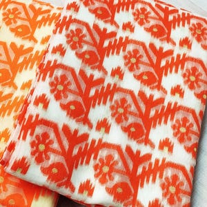 May include: Two folded fabrics with a repeating floral pattern. The design features orange flowers and geometric shapes on a white background. The fabric on the left has a lighter orange hue.