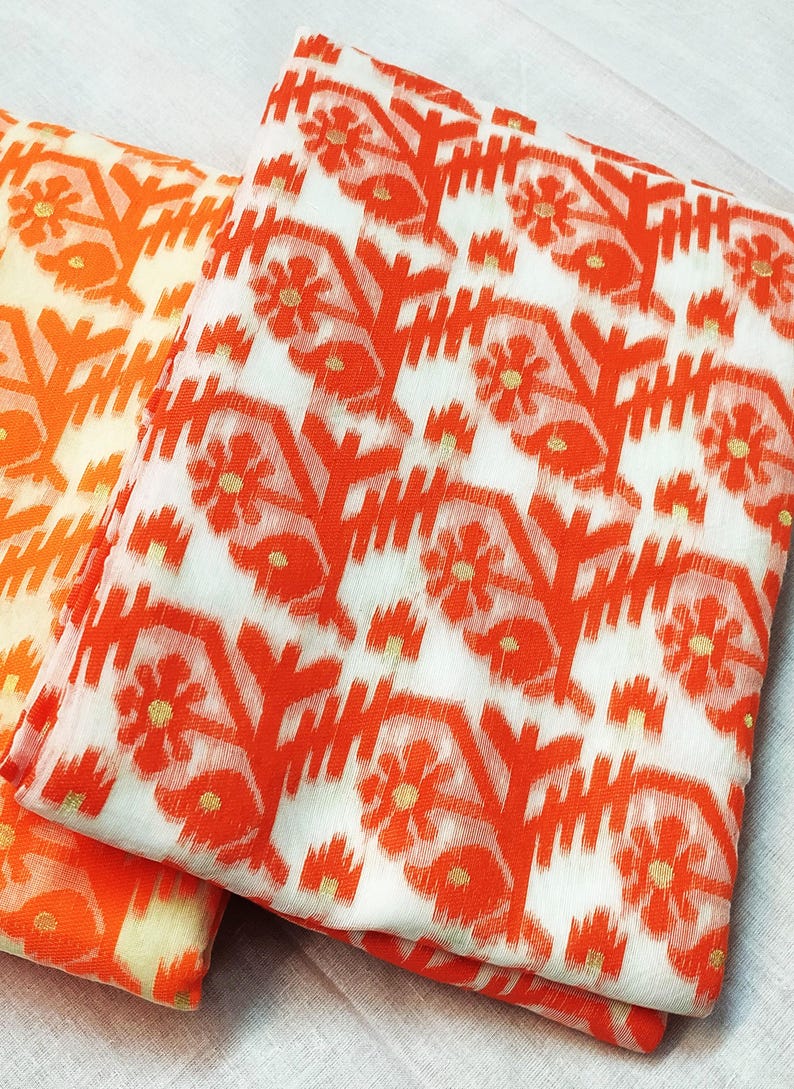 May include: Two folded fabrics with a repeating floral pattern. The design features orange flowers and geometric shapes on a white background. The fabric on the left has a lighter orange hue.