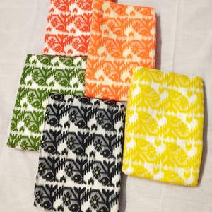 May include: Five folded fabric squares in various colors: orange, red, green, black and white, and yellow. Each square features a repeating floral pattern. The fabrics are arranged on a white surface.