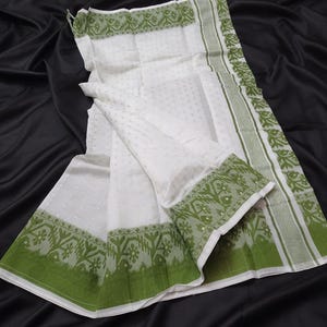 May include: White fabric with a subtle dotted pattern and green borders. The borders feature a repeating floral design and a solid green stripe. The fabric is draped over a black surface.