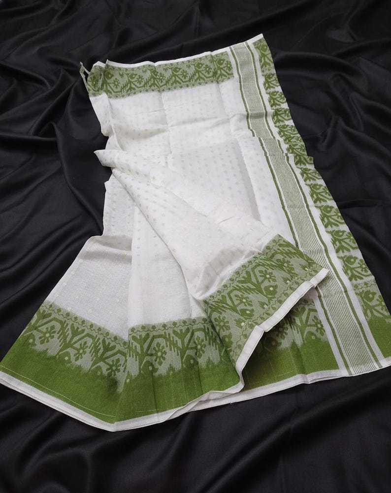 May include: White fabric with a subtle dotted pattern and green borders. The borders feature a repeating floral design and a solid green stripe. The fabric is draped over a black surface.