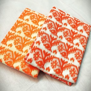 May include: Two folded fabrics with a floral pattern in orange and white. One fabric has an orange background, the other has a white background. The floral design includes stylized flowers and geometric shapes.