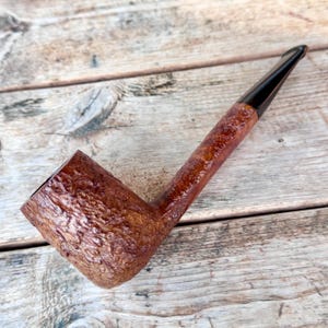 以下が含まれることがあります： 木製の茶色のパイプ。 ざらざらしたボウルと滑らかで暗いステムが特徴です。 パイプはクラシックな曲線デザインです。 木の表面に置かれています。