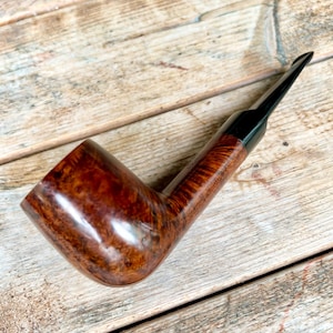 May include: A brown, curved tobacco pipe with a glossy finish. The pipe features a dark, angled mouthpiece and a rounded bowl. The wood grain is visible, showcasing a rich, natural pattern. The pipe rests on a wooden surface.