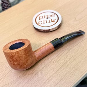 Peut inclure: Une pipe à tabac en bois brun clair avec une finition lisse et polie et un tuyau noir. Derrière la pipe se trouve un disque en bois rond et clair portant le texte pipe club en lettres brunes avec une petite icône de pipe entre les mots.