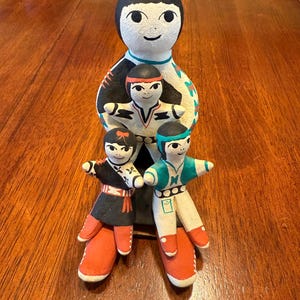 May include: A handcrafted ceramic family figurine. The figures have black hair and painted faces. The adult figure is holding three smaller figures. The figures are wearing colorful clothing, including red, black, and turquoise.