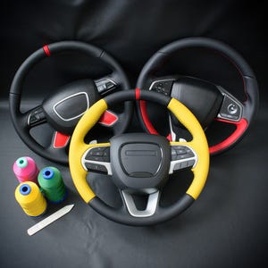 May include: Three black steering wheels with red and yellow accents are displayed against a dark background. One wheel has yellow and black leather, while the others have red accents. Spools of thread and a tool are also visible.
