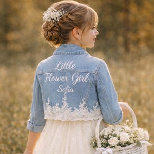 May include: A light blue denim jacket with the words "Little Flower Girl Sofia" embroidered on the back. The jacket has a white lace trim and is worn over a white dress. A white basket filled with white flowers is held in the hand.