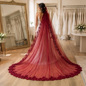 May include: A floor-length, deep red bridal veil featuring intricate dark red floral lace embroidery along the edges. The sheer, flowing tulle fabric extends into a long train, draping over a white, sleeveless gown with a low-cut back design.