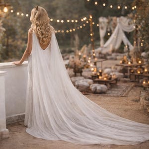 May include: A white wedding dress with a long, flowing train and a sheer cape. The dress has a low back and is worn outdoors. The background features string lights and a decorated outdoor setting, suggesting a wedding or special event.