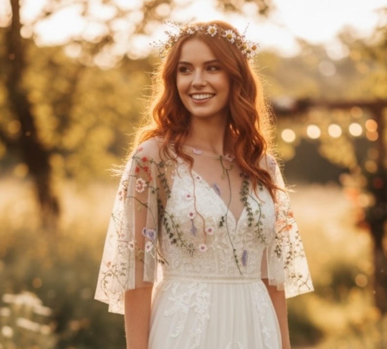 May include: A white dress featuring a sheer overlay with colorful floral embroidery on the sleeves and bodice, paired with a delicate daisy flower crown. The garment has a V-neckline and a fitted waist, showcasing intricate needlework details.