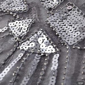 May include: Close-up of a gray fabric embellished with silver sequins and beads. The sequins are arranged in geometric patterns, including triangles and rectangles. The beads are used to create lines and borders, adding texture and dimension to the design.