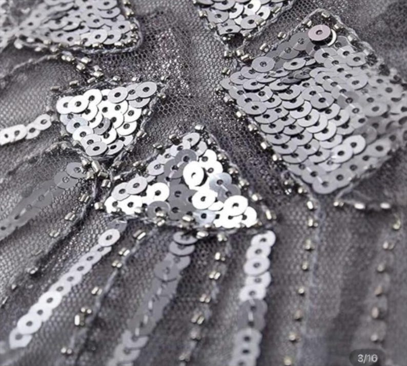 May include: Close-up of a gray fabric embellished with silver sequins and beads. The sequins are arranged in geometric patterns, including triangles and rectangles. The beads are used to create lines and borders, adding texture and dimension to the design.