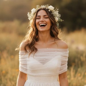 Puede incluir: Un vestido blanco de hombros descubiertos con una capa de tul transparente y fruncido, adornado con peque&ntilde;as perlas blancas espaciadas uniformemente. La prenda se complementa con una corona de rosas blancas y flores de paniculata.