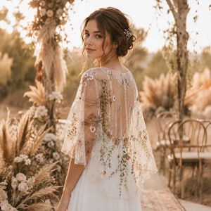 May include: A sheer white bridal capelet featuring delicate, multi-colored floral embroidery with green vines and small pink and white blossoms. The capelet has a wide, flowing silhouette that drapes over the shoulders, worn over a simple white dress.