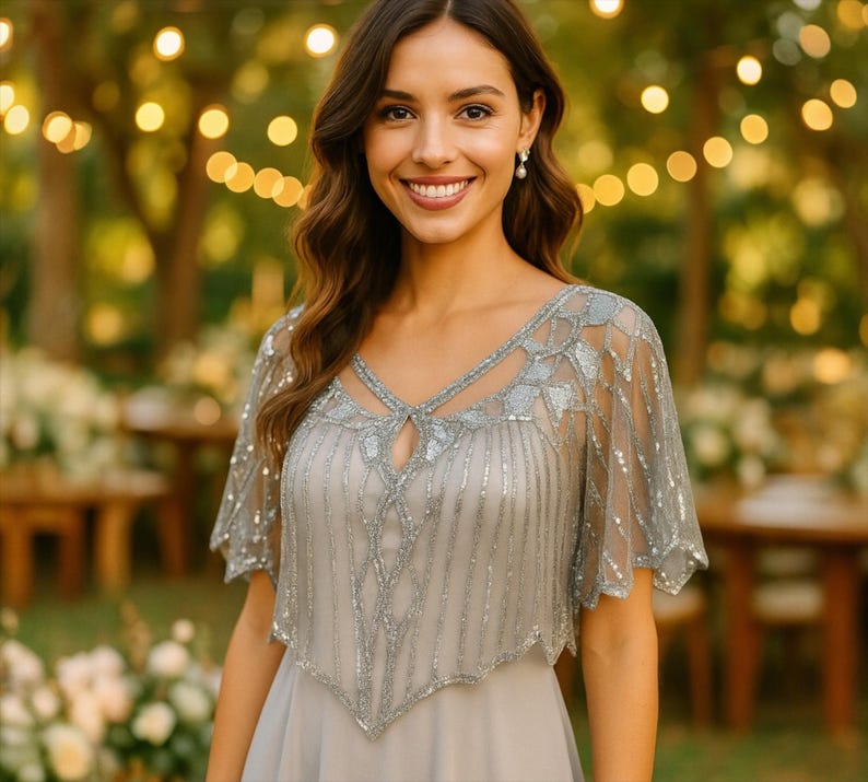 May include: A light gray dress with a beaded overlay. The overlay features intricate silver beading and a cape-like design. The dress has a V-neckline and is worn by a person smiling in an outdoor setting with blurred lights in the background.