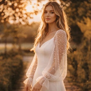 May include: A white wedding dress with a V-neckline and long, sheer sleeves adorned with small, white pearls. The sleeves have ruffled cuffs. The dress is worn by a person with long, blonde hair and a floral hairpiece, set against a blurred outdoor background.
