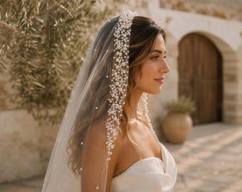May include: A sheer, floor-length bridal veil featuring a dense, cascading arrangement of white pearls along the crown and edges, transitioning into a sparse scattering of individual pearls across the length of the lightweight tulle fabric.
