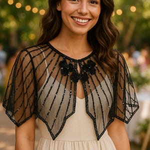 May include: Black sheer capelet with a geometric design and sequin accents. The capelet features a floral embellishment at the neckline and is worn over a cream-colored dress. Perfect for formal events or adding a touch of elegance.