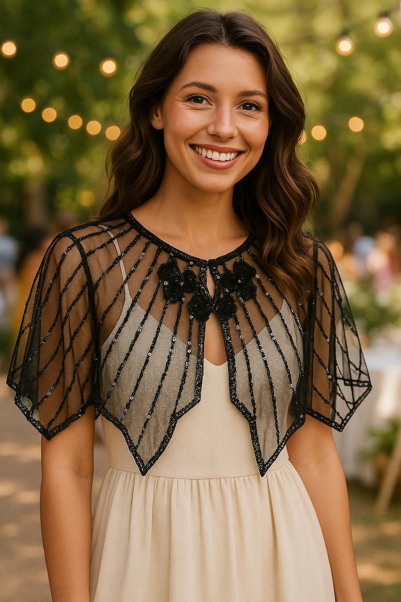 May include: Black sheer capelet with a geometric design and sequin accents. The capelet features a floral embellishment at the neckline and is worn over a cream-colored dress. Perfect for formal events or adding a touch of elegance.