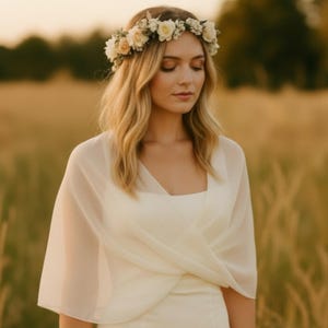 May include: A woman wearing a white dress, a sheer shawl, and a floral crown. The dress has a square neckline. The shawl is draped over the shoulders. The floral crown is made of white and yellow flowers. The background is a field of tall grass.