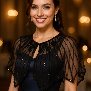 May include: A woman wearing a black sequined cape over a navy blue dress. The cape has a sheer, delicate design with vertical and diagonal lines of sequins. The dress has a V-neckline and a flowing skirt. The background is blurred with warm, golden tones.