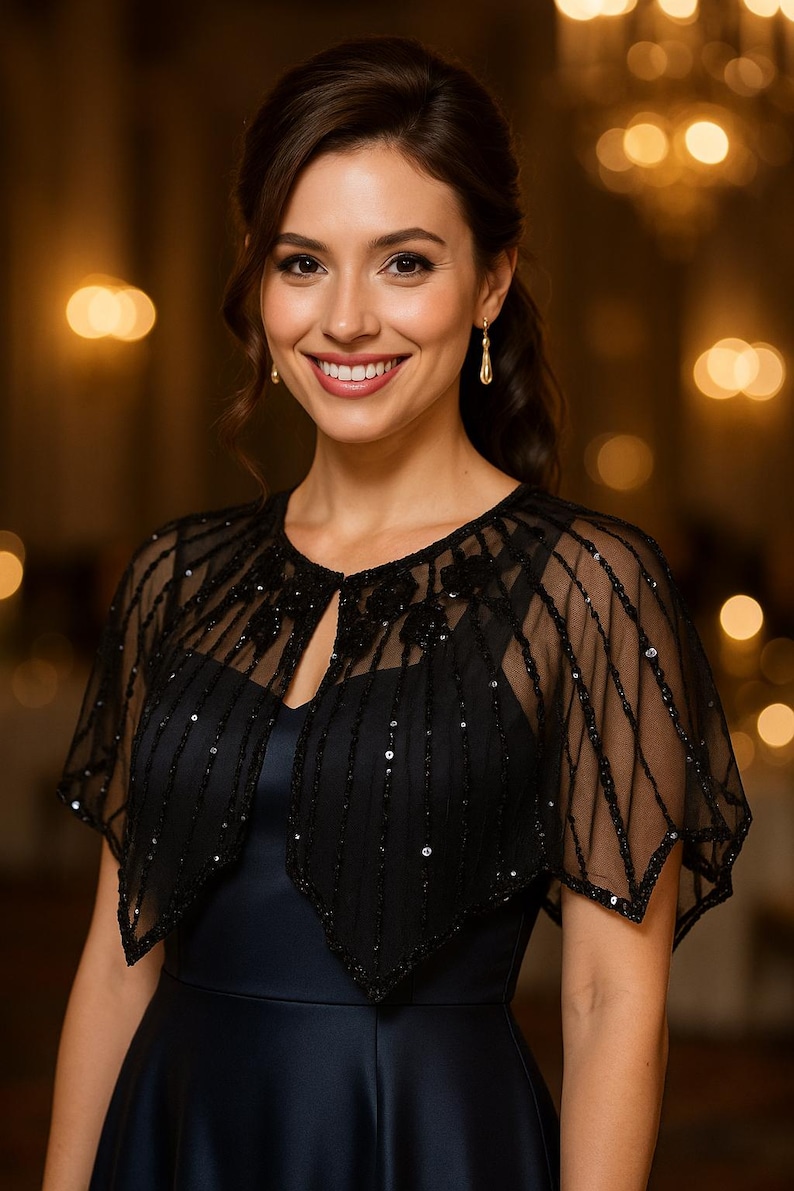 May include: A woman wearing a black sequined cape over a navy blue dress. The cape has a sheer, delicate design with vertical and diagonal lines of sequins. The dress has a V-neckline and a flowing skirt. The background is blurred with warm, golden tones.