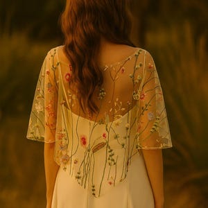 May include: A sheer, short-sleeved capelet featuring colorful floral embroidery with green stems and various blossoms, worn over a solid white dress. A matching floral crown with small white flowers rests on the hair, completing the ensemble.