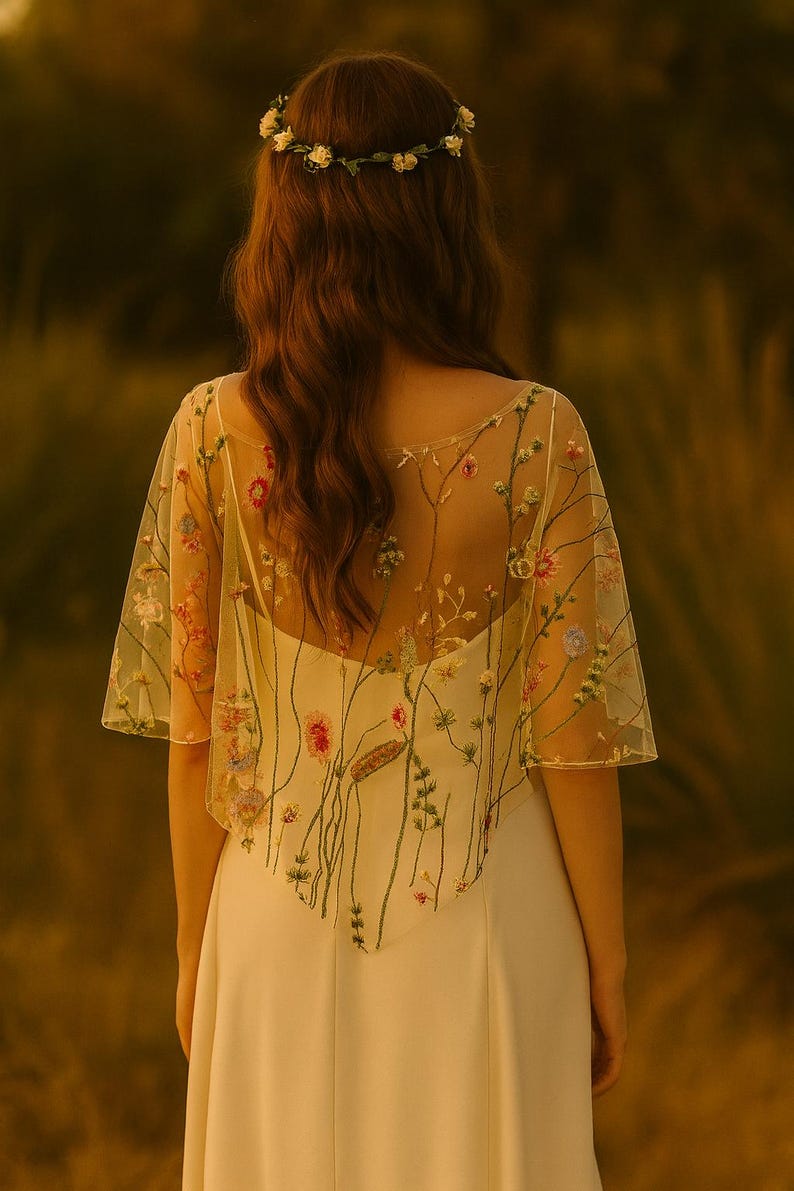 May include: A sheer, short-sleeved capelet featuring colorful floral embroidery with green stems and various blossoms, worn over a solid white dress. A matching floral crown with small white flowers rests on the hair, completing the ensemble.