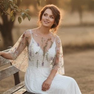 May include: A white floor-length dress featuring a sheer overlay with delicate floral embroidery. The bodice has a V-neckline and short, flowing sleeves. The embroidery includes small green leaves and purple and white flowers scattered across the fabric.