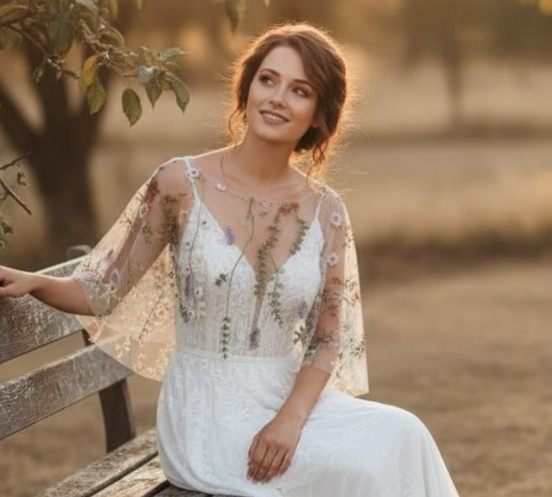 May include: A white floor-length dress featuring a sheer overlay with delicate floral embroidery. The bodice has a V-neckline and short, flowing sleeves. The embroidery includes small green leaves and purple and white flowers scattered across the fabric.