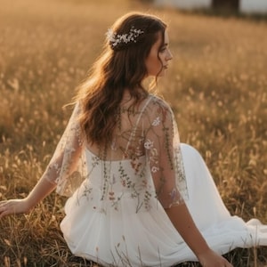 May include: A white dress featuring a sheer overlay capelet adorned with delicate, multi-colored floral embroidery. The garment has short, flared sleeves and a flowing skirt. A silver-toned hair accessory with small white flowers is worn.