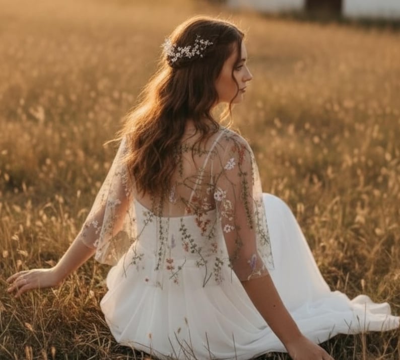 May include: A white dress featuring a sheer overlay capelet adorned with delicate, multi-colored floral embroidery. The garment has short, flared sleeves and a flowing skirt. A silver-toned hair accessory with small white flowers is worn.