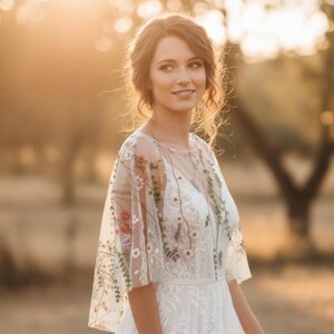 May include: A white sheer bridal capelet featuring delicate, multi-colored floral embroidery with green vines and small blossoms. The garment has a wide, flowing silhouette with short sleeves and a round neckline, worn over a white lace dress.