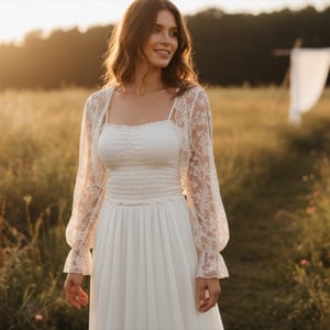 May include: A white wedding dress with a fitted bodice and flowing skirt. A sheer, floral lace bolero jacket with long sleeves is worn over the dress. The image is taken outdoors in a field with sunlight.