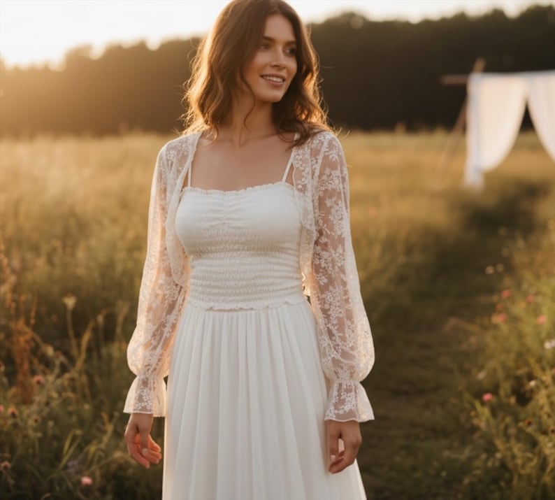 May include: A white wedding dress with a fitted bodice and flowing skirt. A sheer, floral lace bolero jacket with long sleeves is worn over the dress. The image is taken outdoors in a field with sunlight.