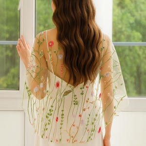May include: A sheer, off-white capelet featuring delicate, multi-colored floral embroidery with thin green stems and small red, pink, and blue blossoms. The lightweight, translucent fabric drapes over the shoulders, showcasing intricate botanical details.