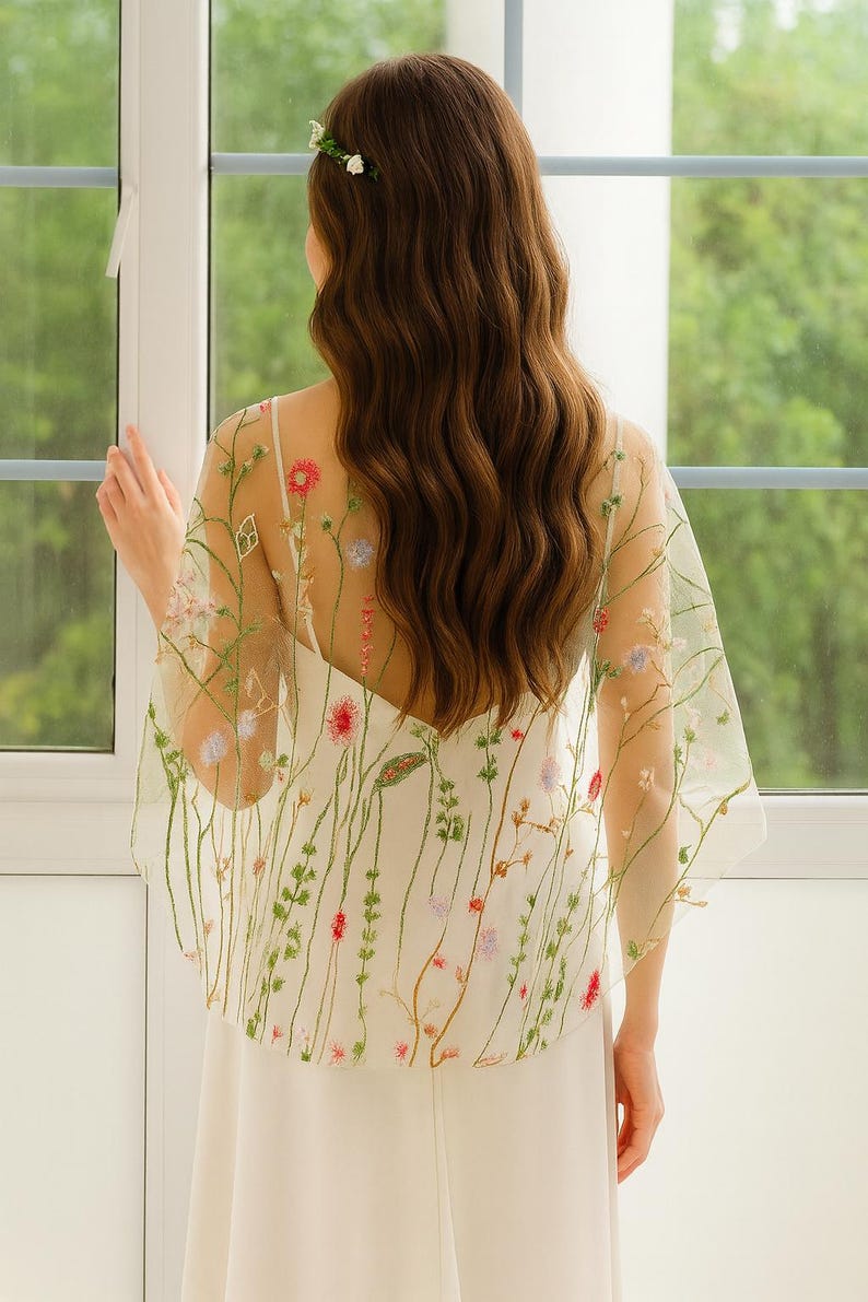 May include: A sheer, off-white capelet featuring delicate, multi-colored floral embroidery with thin green stems and small red, pink, and blue blossoms. The lightweight, translucent fabric drapes over the shoulders, showcasing intricate botanical details.