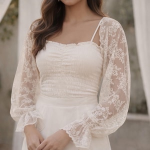 May include: Ivory-colored bridal top and skirt. The top features a square neckline and a ruched bodice. Delicate lace sleeves with floral patterns and flared cuffs add a romantic touch. The skirt is a simple, flowing design. Perfect for a wedding or special occasion.