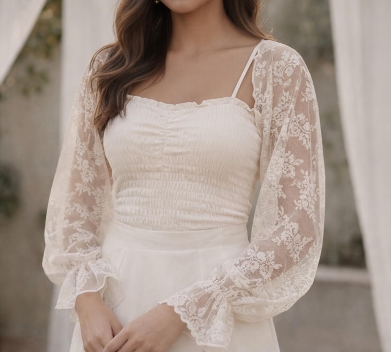 May include: Ivory-colored bridal top and skirt. The top features a square neckline and a ruched bodice. Delicate lace sleeves with floral patterns and flared cuffs add a romantic touch. The skirt is a simple, flowing design. Perfect for a wedding or special occasion.