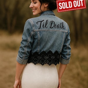 May include: A blue denim jacket featuring black lace trim along the bottom hem and the text Til Death embroidered in black script across the upper back. The jacket has long sleeves with folded cuffs and a classic collar, worn over a white dress.